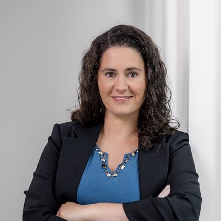 Eine Person in einem blauen Oberteil und einem schwarzen Blazer steht mit verschränkten Armen vor einer weißen Wand. Die Atmosphäre wirkt professionell.